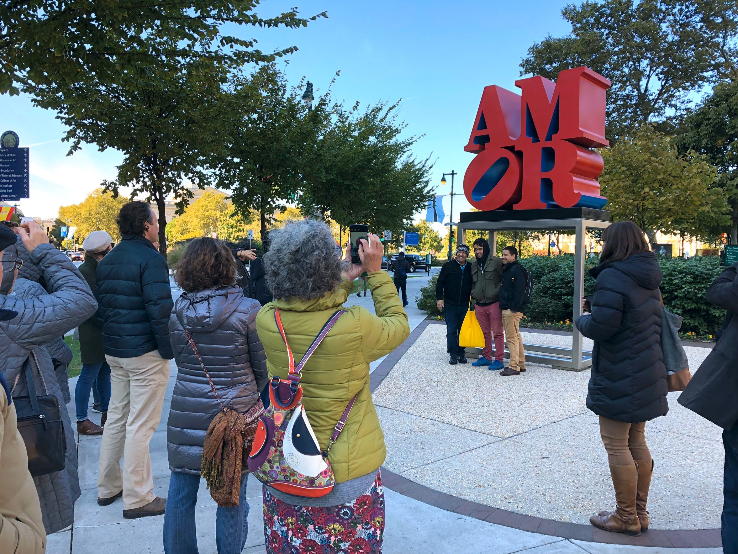 A group tour on the Benjamin Franklin Parkway stops to take photos at Robert Indiana's "AMOR" scupture - bright red staked letters that spell A-M-O-R on a tall metal frame base