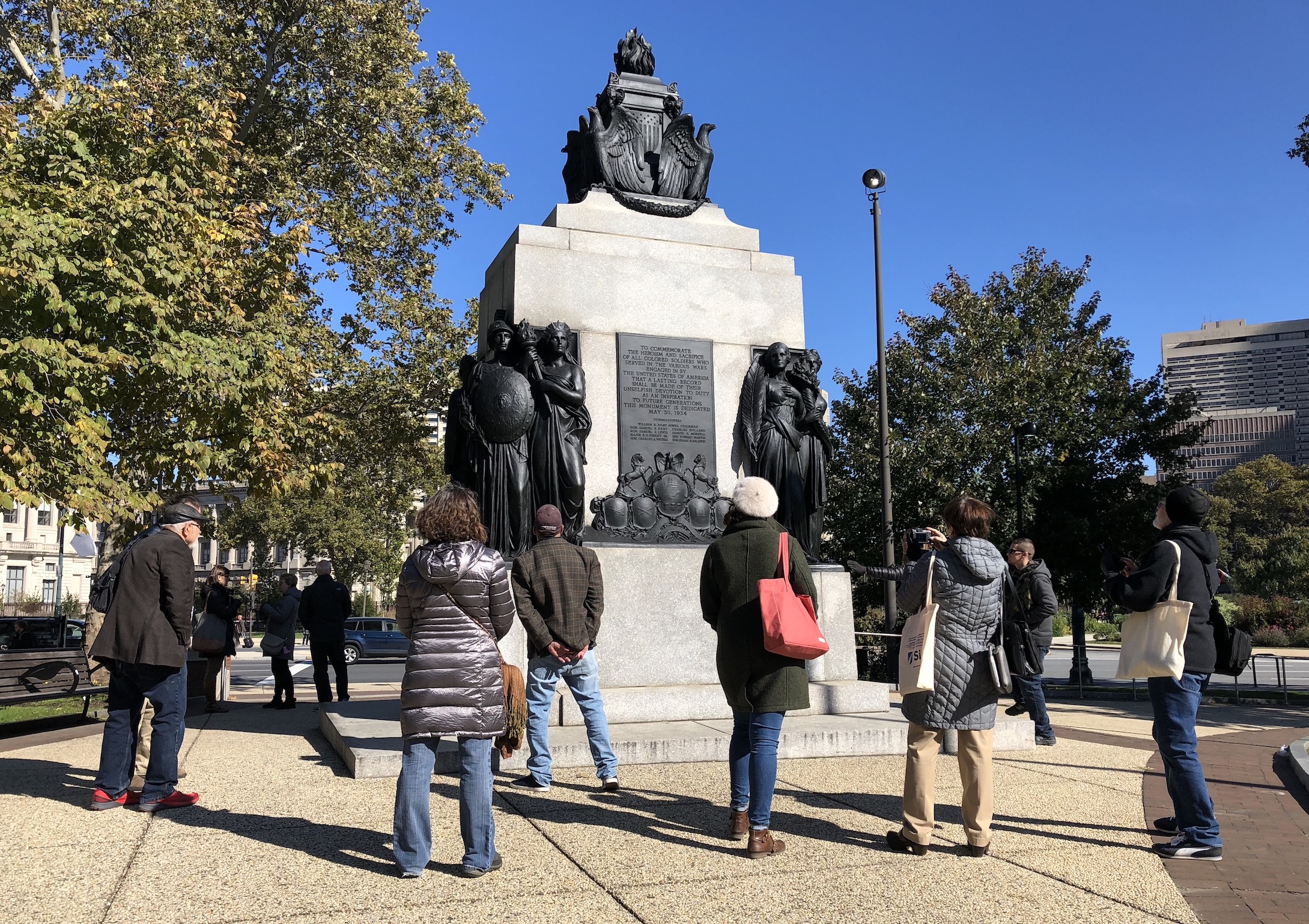 A group on an outdoor sculpture tour around the All Wars Memorial, looking up at the memorial and taking photos on a sunny fall day on the Parkway.