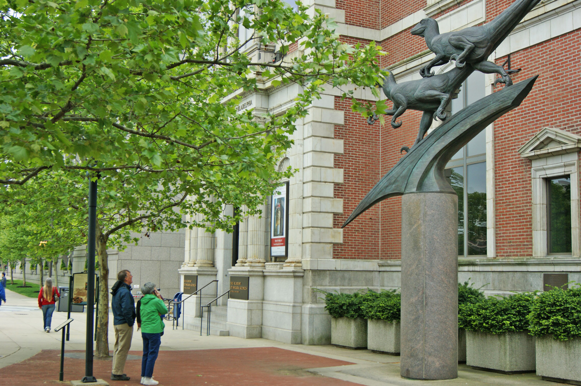 Two people look up toward the bronze Deinonychus dinosaur sculptures in front of the Academy of Natural Sciences on the Benjamin Franklin Parkway