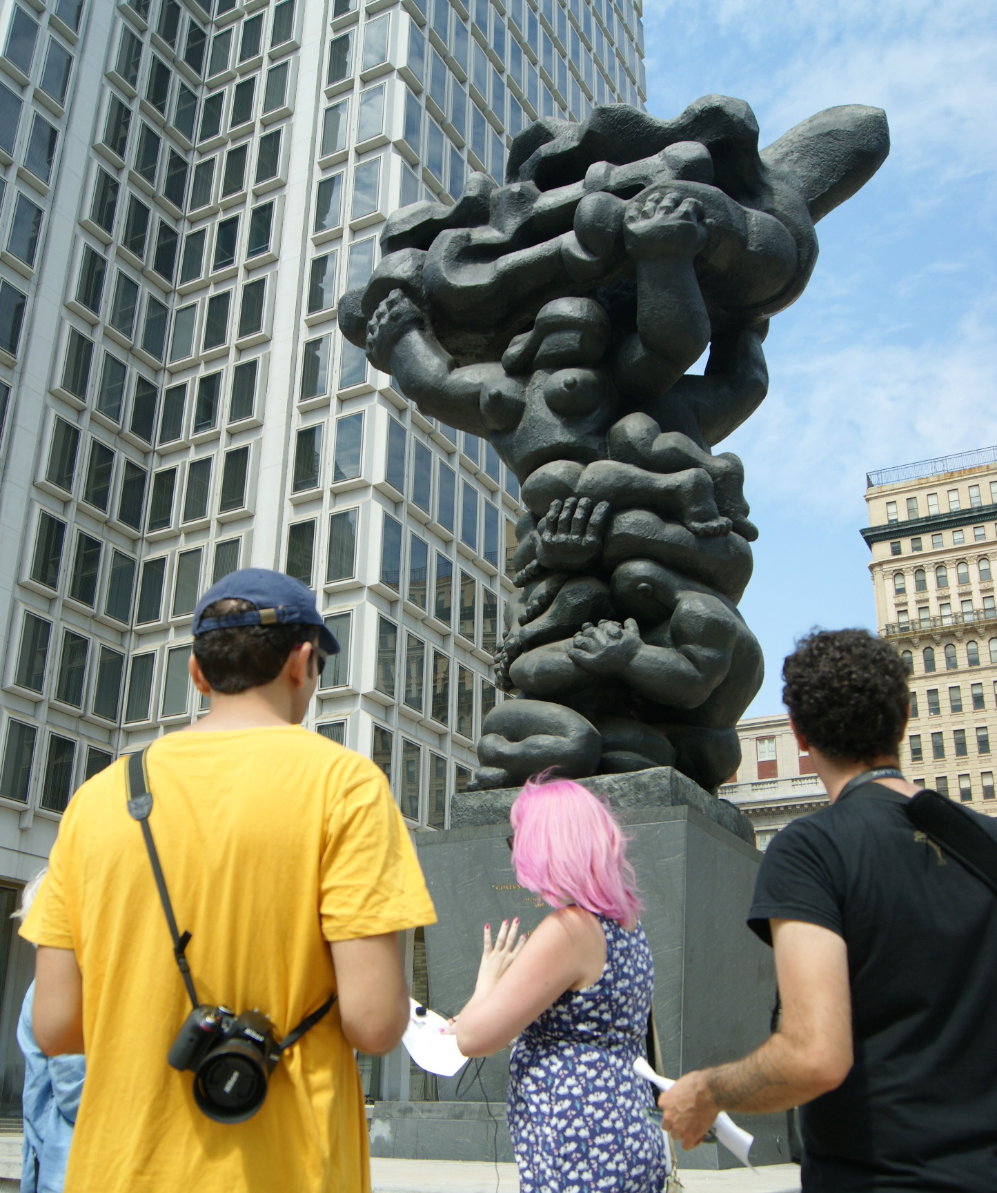 Three people participating in a public art photography workshop at the massive bronze Jacques Lipchitz "Government of the People" sculpture in Center City Philadelphia. The instructor is a woman with pink hair in the center and she is talking about the sculpture. Two men with their cameras are listening.