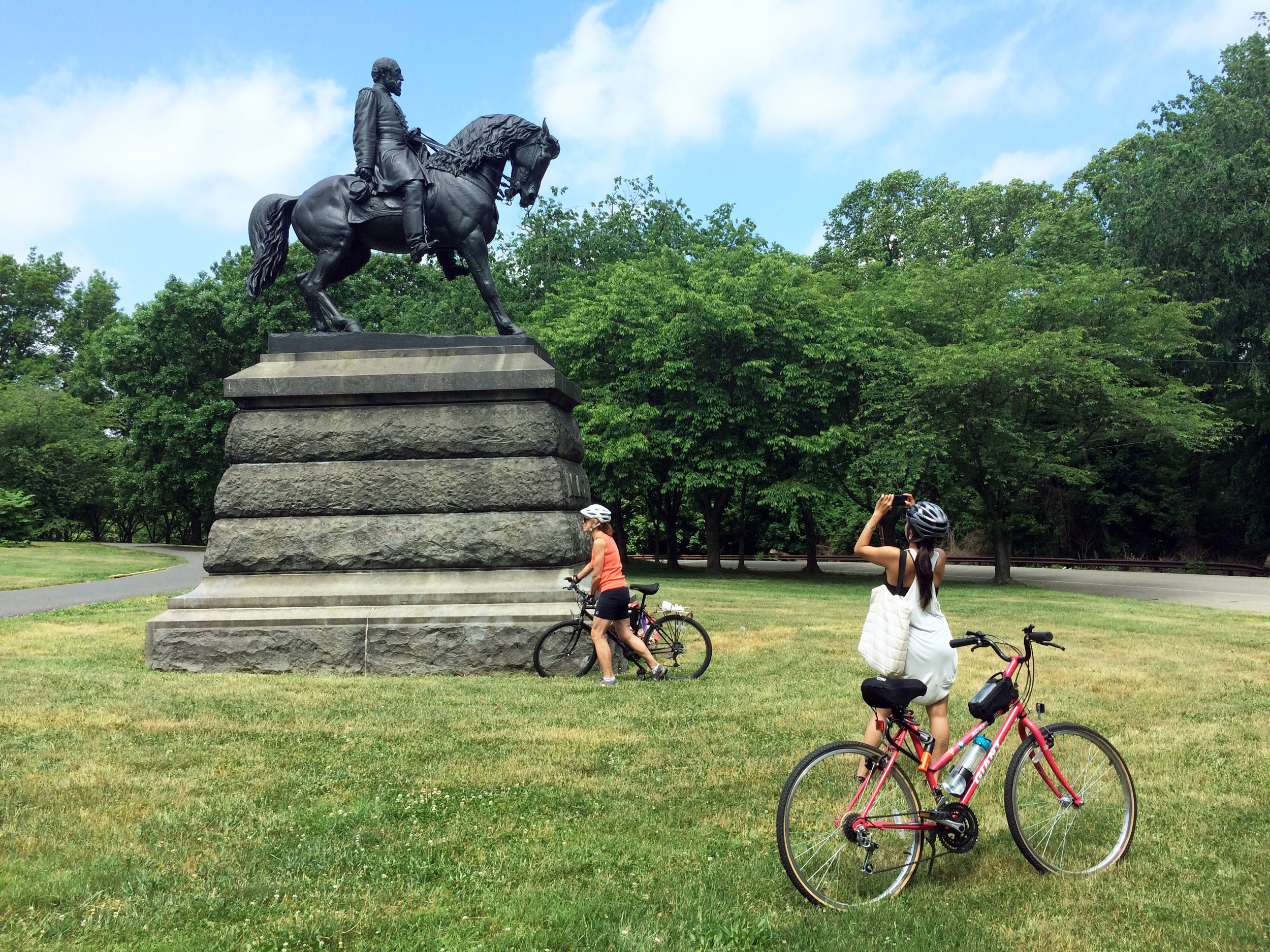 Two female cyclists at the Meade bronze equestrian in Fairmount Park. One of the cyclists stops to take a picture of the monument, which stands on a tall stone base.