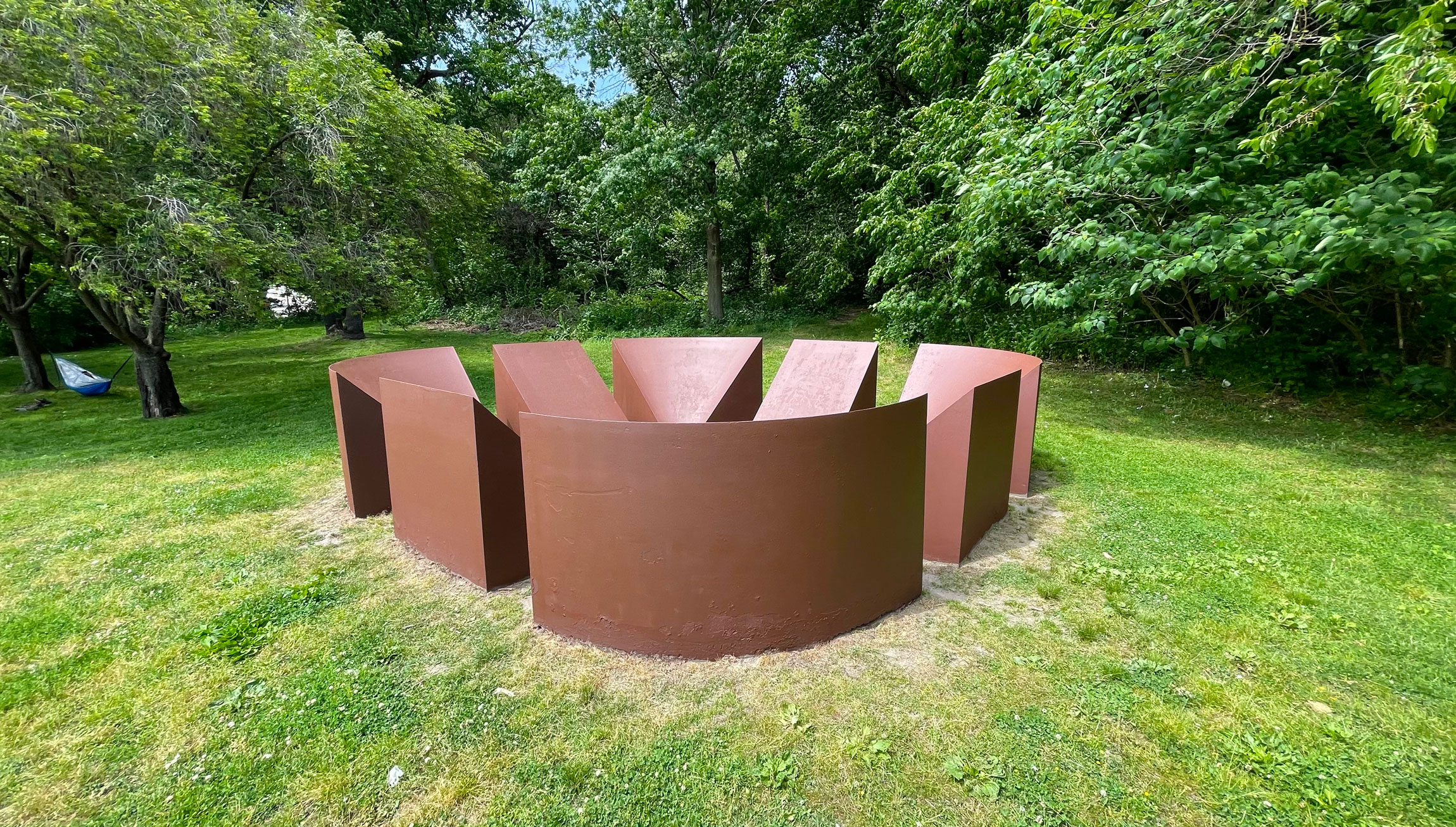 Robert Morris' corten steel reddish brown sculpture with green grass and trees. They are eight geometric wedge forms arranged on the ground in a circular square