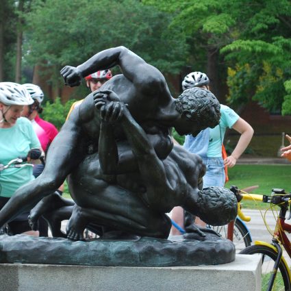 Cyclists on a public art bike tour make a stop at the bronze "Wrestlers" sculpture in Fairmount Park. The tour leader is gesturing at and talking about the artwork.