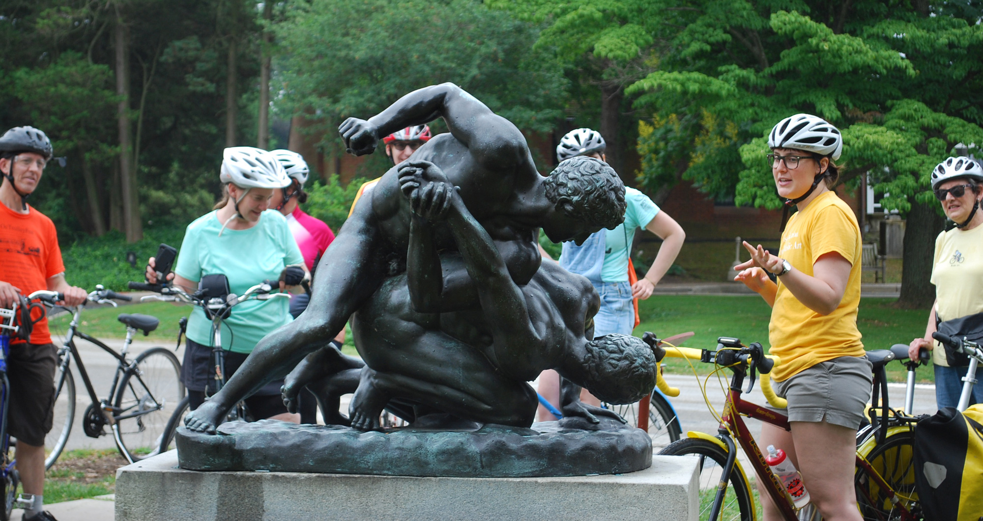 Cyclists on a public art bike tour make a stop at the bronze "Wrestlers" sculpture in Fairmount Park. The tour leader is gesturing at and talking about the artwork.