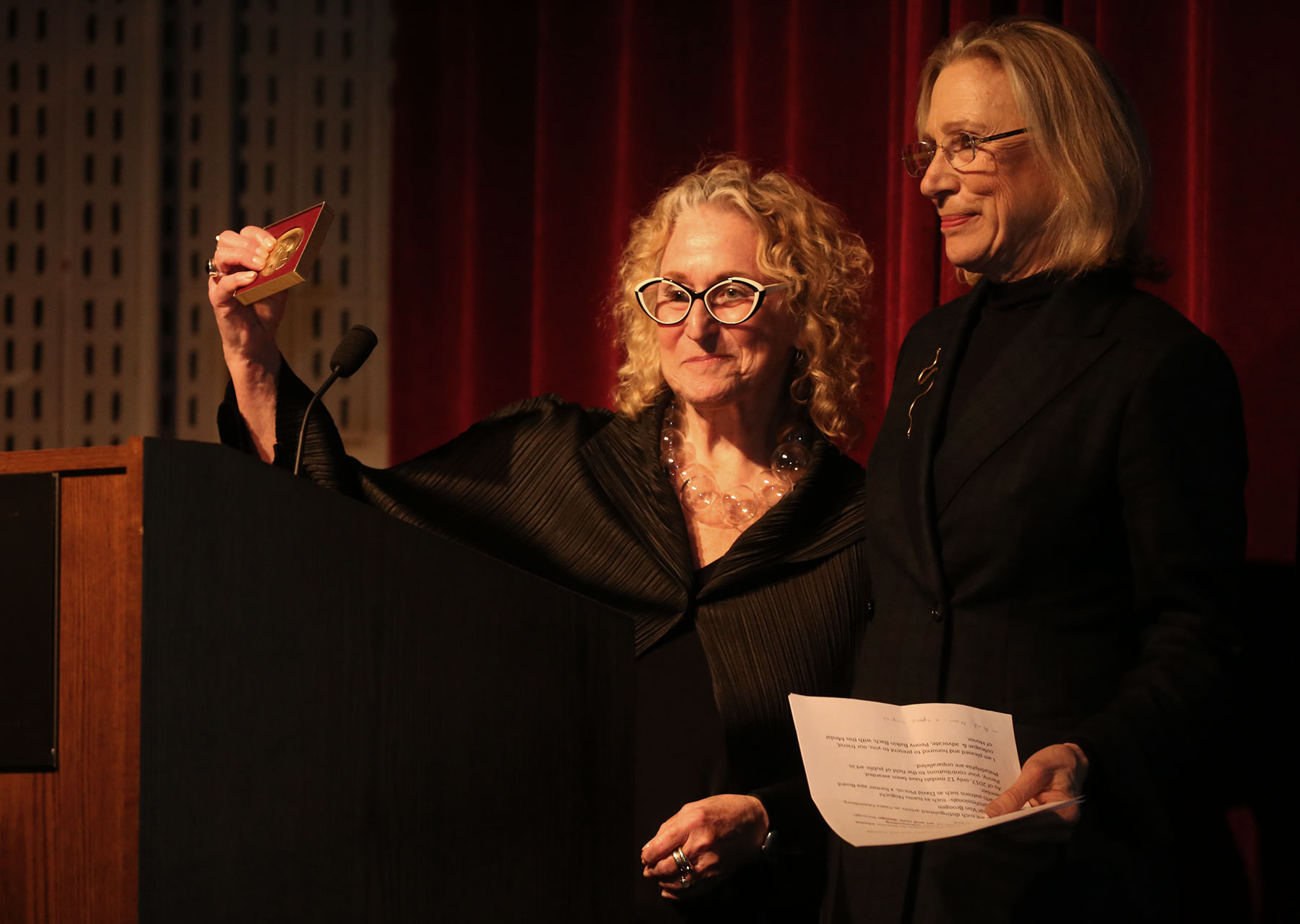 Penny Balkin Bach, longtime aPA Executive Director, holds up the medal of honor she has just received. She is standing next to Board President Barbara Aronson. Both women, smiling, have blonde hair are wearing black clothes, and behind them are red stage curtains. It is the Association for Public Art's Annual Meeting event at Moore College of Art and Design in Philadelphia, PA.