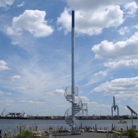 A mast-like object with a spiral staircase wrapped around its base with the river in the background and a blue sky with large white clouds