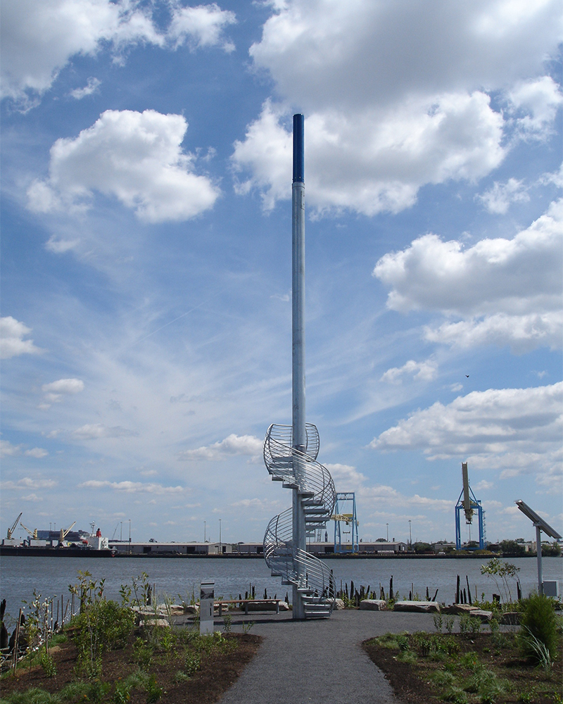 a mast-like object with a spiral staircase wrapped around its base with the river in the background and a blue sky with large white clouds