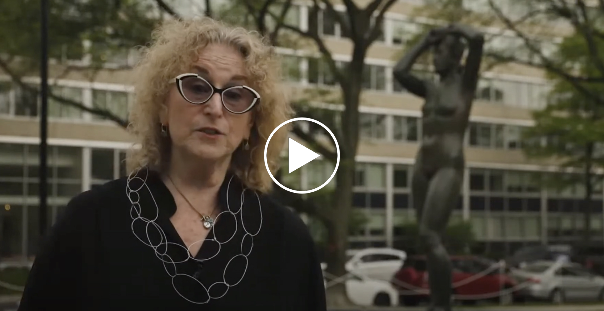 aPA's Executive Director on site with the Maja sculpture, giving a talk about the artwork's history. Penny has blonde curly hair, black and white cat eye glasses, a black top and a necklace of thin white circles
