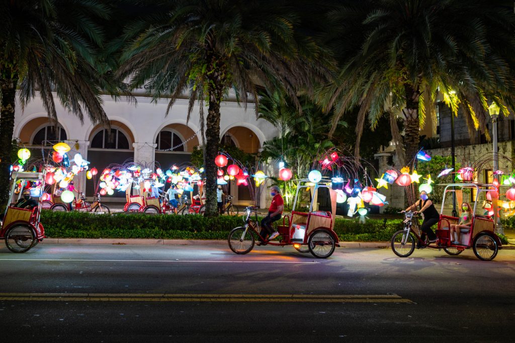 In the foreground and background, pedicabs adorned with glowing handmade lanterns glide along the street in Florida with palm trees in the middle ground.