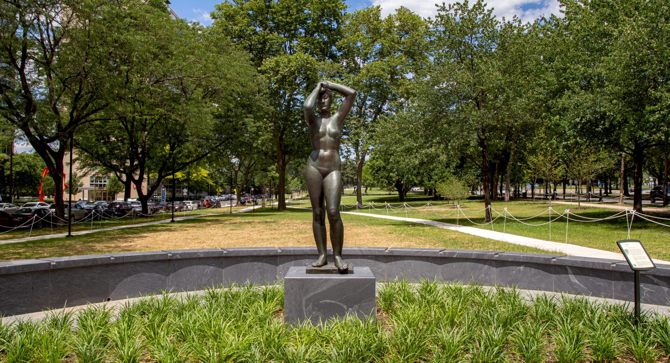 The Gerhard Marcks "Maja" sculpture – figurative bronze nude female with arms overhead – in the new "Maja Park" on the Parkway on a sunny June day. The sculpture stands tall on a gray granite base, surrounded by green Liriope plants.