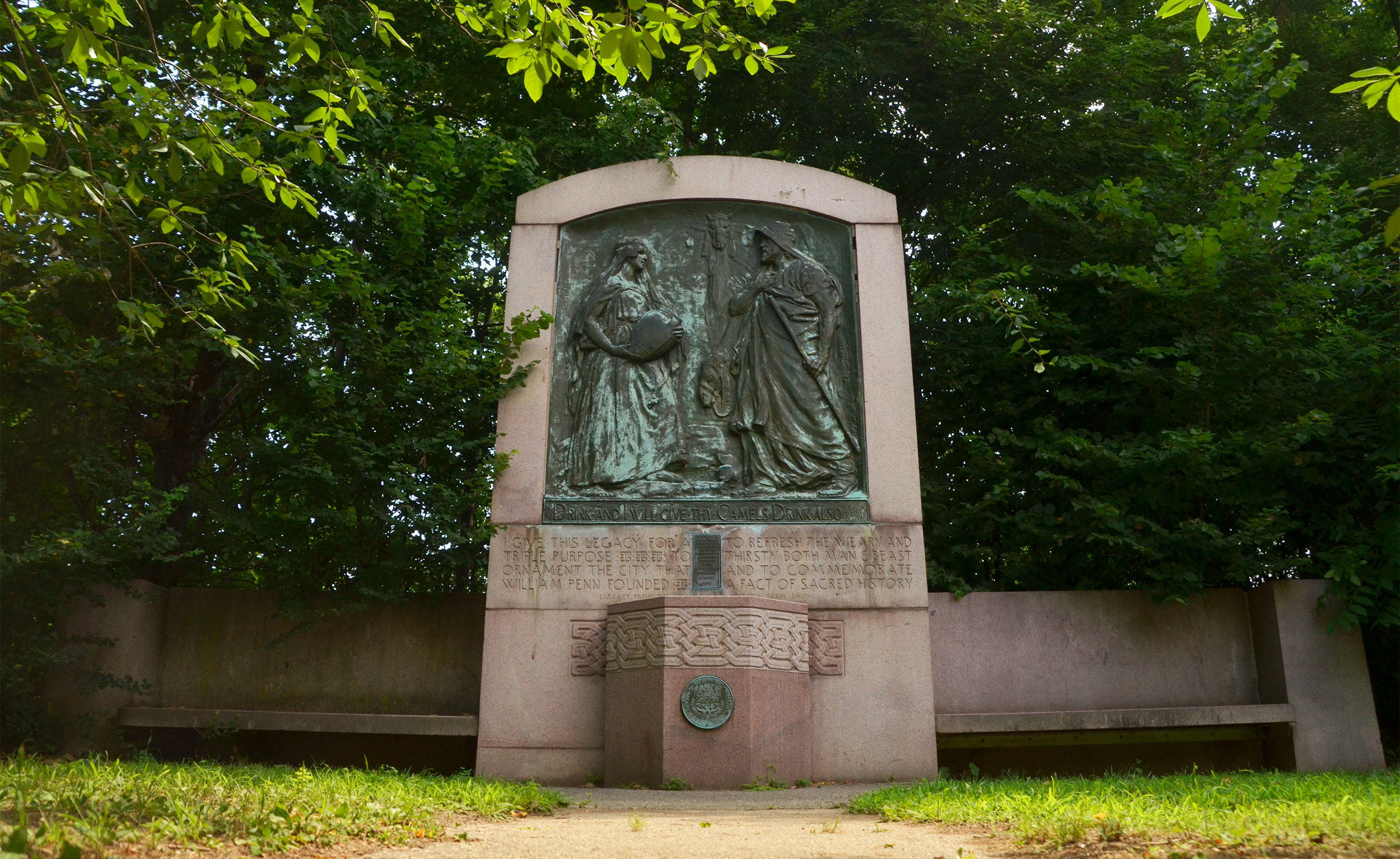 Full view of the weathered bronze relief in Fairmount Park that depicts a biblical scene: Rebecca offering water to a man and his camel. The bronze relief is set in a granite structure with seating and an old water fountain.