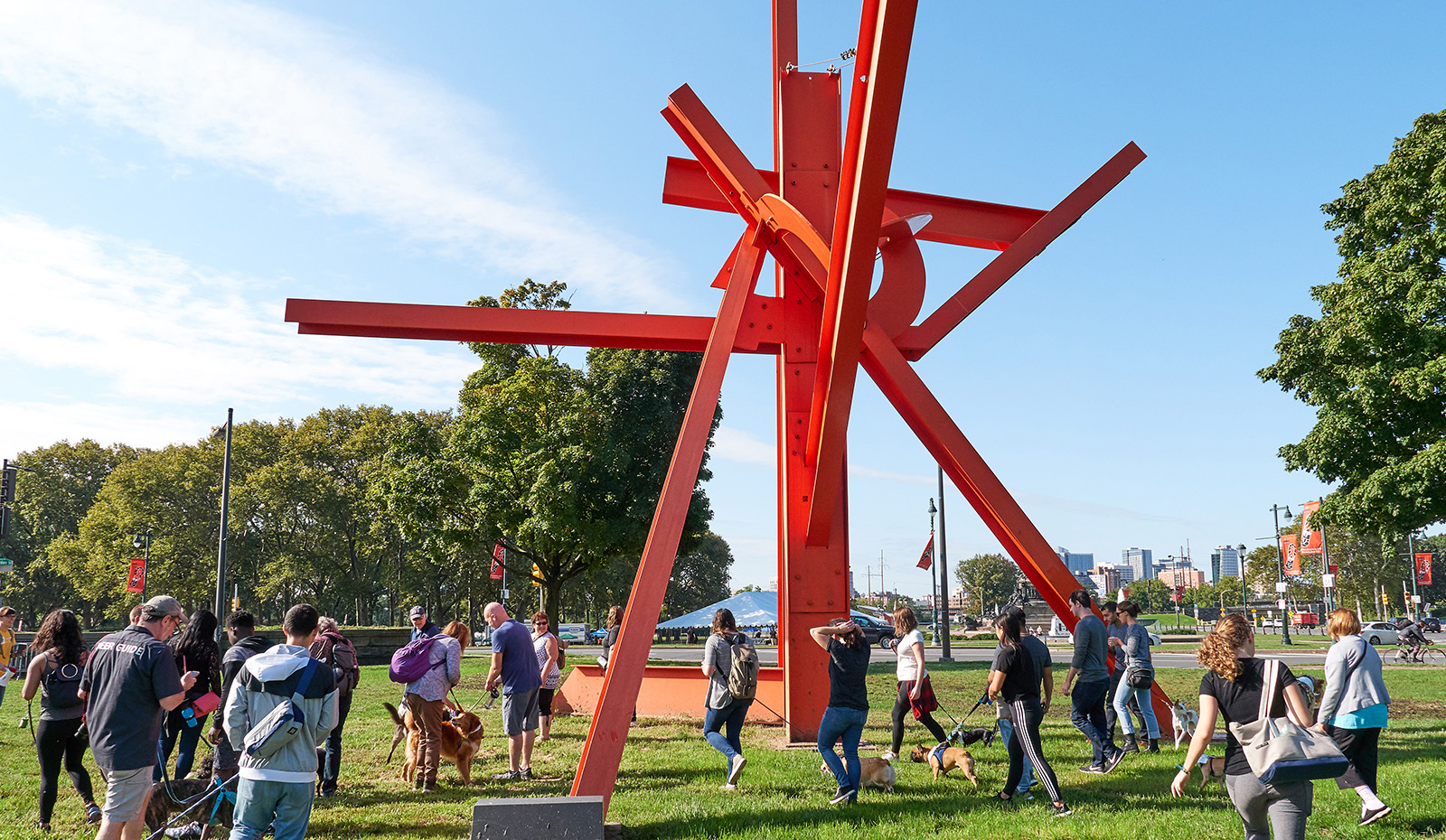Bright red abstract i-beam sculpture on a bright sunny day with people walking their dogs around the sculpture. Artist is Mark di Suvero.