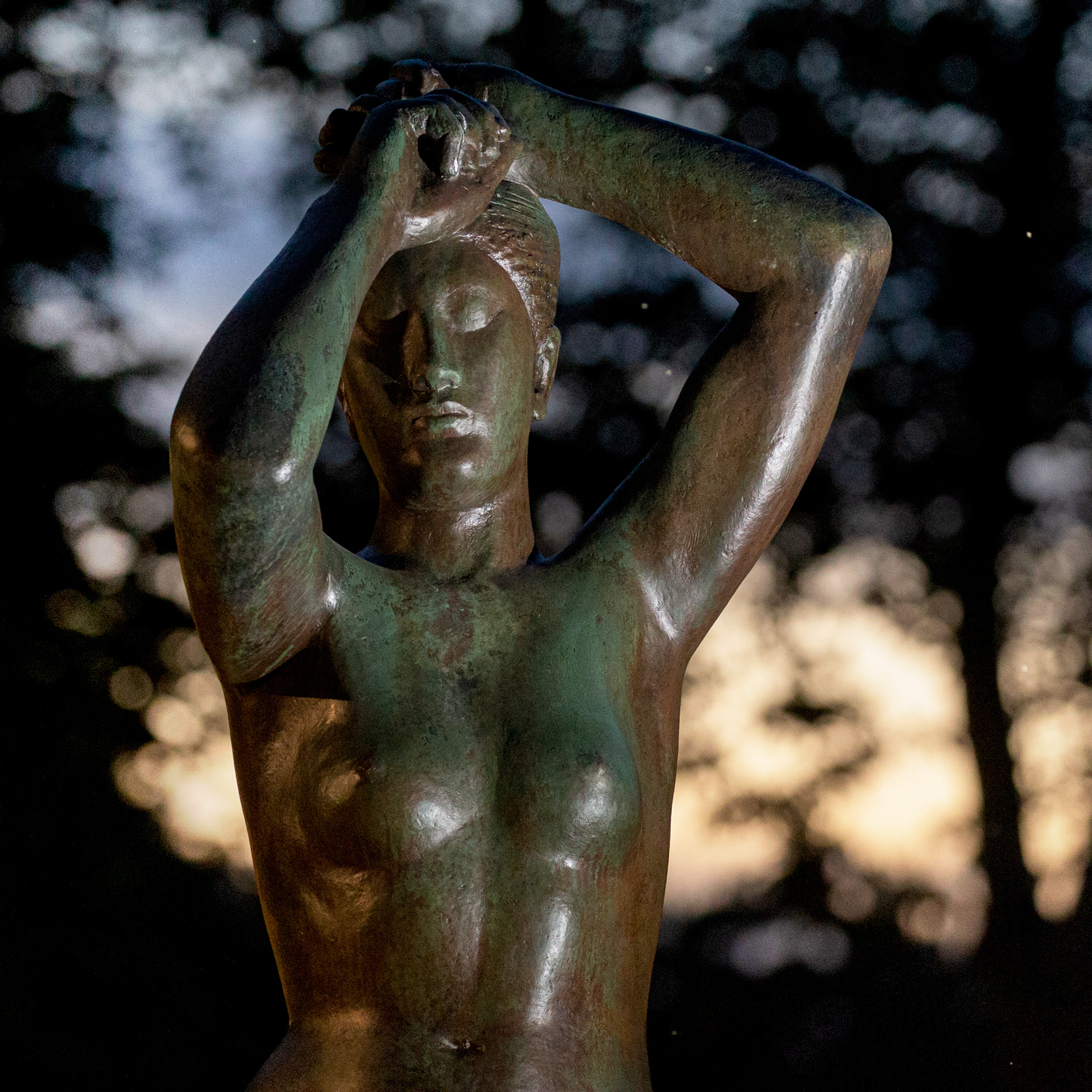 A nighttime view of a bronze nude female figure with her arms stretched over her head