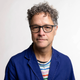 Headshot of an artist, who is a man in his 50s with curly gray hair, eyeglasses, a dark blue jacket, and a multi-color striped shirt.