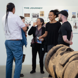 A group of five people smiling and talking at a gallery exhibition with a First Nations carved wood house post in view.