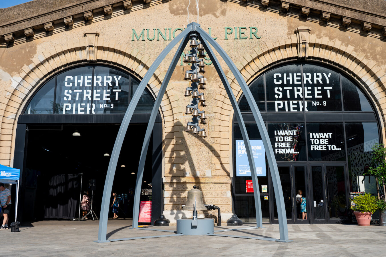 A large grey arch structure – a bell tower – supports a vertical cluster of 32 brass bells. It stands in front of a light-colored building with arched windows, displaying text that reads 'MUNICIPAL PIER No. 9' and 'CHERRY STREET PIER No. 9'.